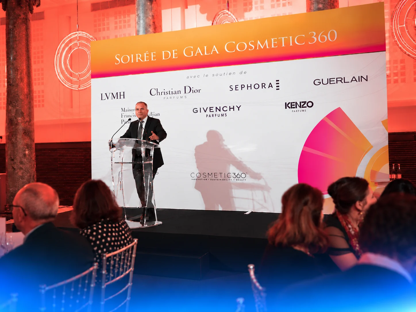 Talare vid en genomskinlig talarstol på Cosmetic 360:s galakväll i Paris, med sittande deltagare och kosmetikvarumärken på scenens bakgrund.